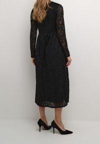 Svart spetsklänning med långa ärmar, en figurnära överdel och en utsvängd kjol. Detaljerat blommönster och lager av textur. Buren med svarta klackar.