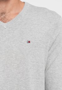 Pilkas V formos megztinis, pagamintas iš minkštos medžiagos, su mažu, raudonos, baltos ir mėlynos spalvų siuvinėtu logotipu kairėje krūtinės pusėje.