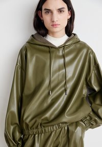 Hoodie en simili cuir vert olive avec cordon de serrage, taille élastique et texture lisse et brillante. Détail de poche avant visible.