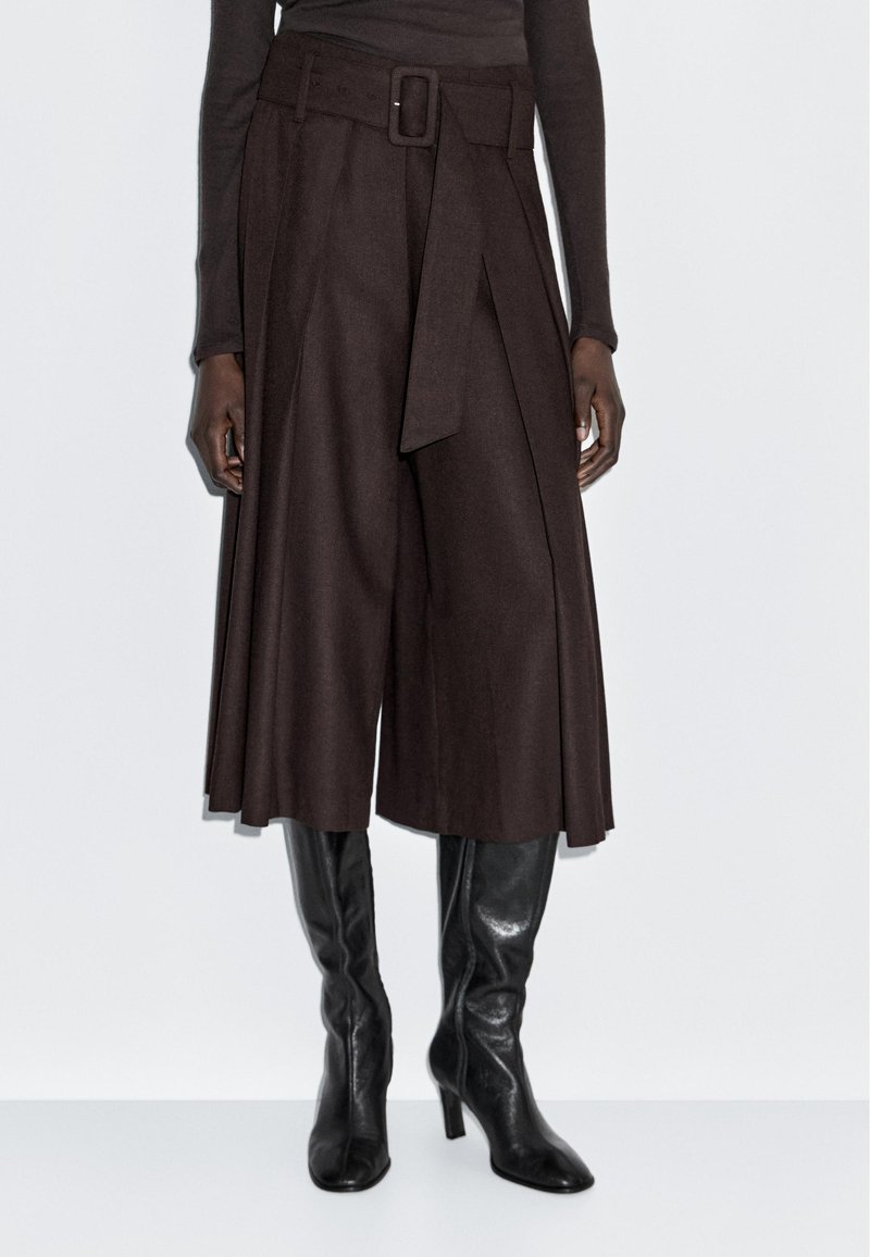 Culottes amples marron foncé avec une ceinture assortie, portés avec des bottes en cuir noires à talons hauts arriver au genou et un haut ajusté à manches longues.