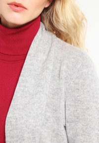 Šedý pletený cardigan s měkkou texturou, otevřen�ým předním designem a pod ním červený rolák. Důraz na vrstvení a neutrální barvy.
