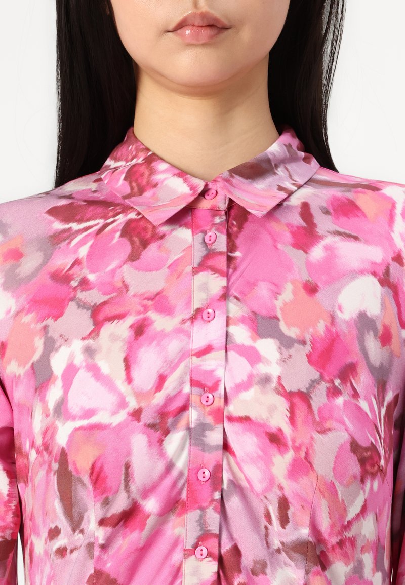 Różowa kwiatowa bluzka z kołnierzykiem, posiadająca zapięcie na guziki, wykonaną z lekkiego materiału oraz o żywych wzorach w stylu akwareli w różnych odcieniach różu.