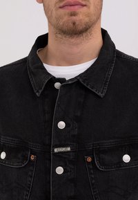Sort denimjakke med krave, sølvfarvede trykknapper, to brystlommer og et mærke på venstre side.