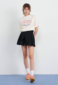 Crème Nike T-shirt met rode tekst en logo, gecombineerd met een zwarte geplooide rok. Model draagt witte sokken en oranje sneakers. Effen achtergrond.