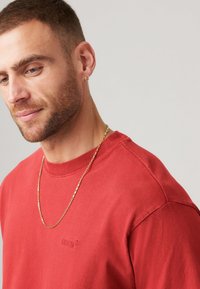 Rød bomulds t-shirt med rund halsudskæring og korte ærmer, prydet med et diskret præget logo. En guldkæde halskæde tilføjer detaljer.