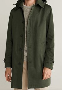 Oliivinvihreä trenssitakki, jossa napit edessä, olkatoppa ja säädettävät hihansuut. Päällä vaaleanharmaa neulepaita. Sileä pinta.