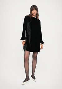 Black velvet mini dress with long bell sleeves and white lace trim at the neckline. Model wears sheer black tights and white heeled mules.