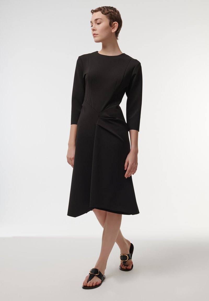 Vestido negro con mangas tres cuartos, que presenta un corsé ajustado, dobladillo asimétrico y detalles drapeados. Modelo llevando sandalias negras con un acento dorado.