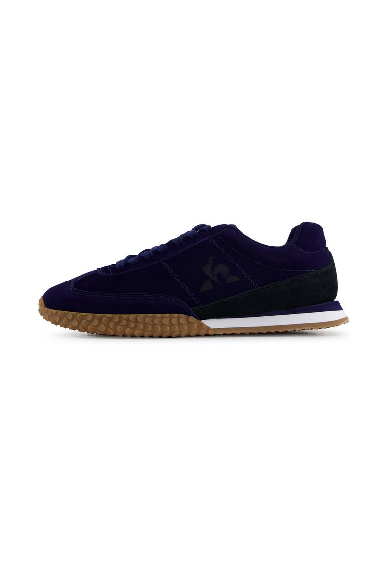 Sneaker low-top con tomaia in suede nero e viola scuro, suola texturizzata marrone e logo nero laterale, mostrato in profilo su sfondo bianco.
