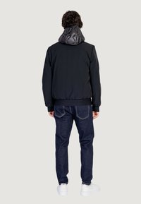 Giubbotto bomber nero con cappuccio trapuntato, polsini e orlo a costine; abbinato a jeans scuri e sneakers bianche. Dettaglio del tessuto testurizzato visibile.