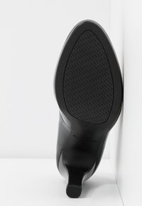 Botte noire à talon avec semelle texturée et bout pointu, positionnée avec la semelle contre un coin mur et sol blancs.