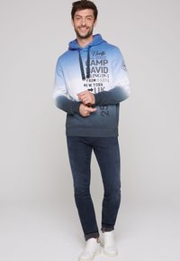 Felpa con cappuccio caratterizzata da una sfumatura che va dal blu chiaro al teal scuro, con testo stampato e dettagli. Abbinata a jeans scuri e sneaker bianche.