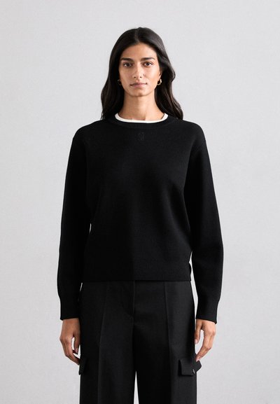 Pull en tricot noir avec un col rond, des poignets et un ourlet côtelés. Présente un logo discret sur la poitrine et est porté avec un pantalon noir.