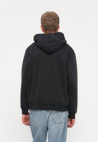Man i svart hoodie och ljusblå jeans, sedd bakifrån mot en enfärgad vit bakgrund.