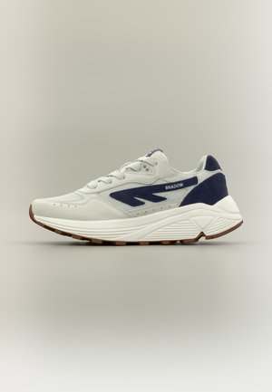 Witte en marineblauwe sportieve sneaker met dikke zool, mesh- en suède materialen, en "SHADOW" tekst aan de zijkant.