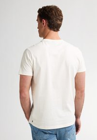 Camiseta de color crema de manga corta con cuello redondo, hecha de tela suave, que presenta una textura lisa y un pequeño logo de la marca en el dobladillo.