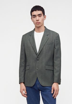 Jeune homme portant un blazer gris à chevrons par-dessus un t-shirt blanc et un jean bleu, debout devant un fond blanc uni.
