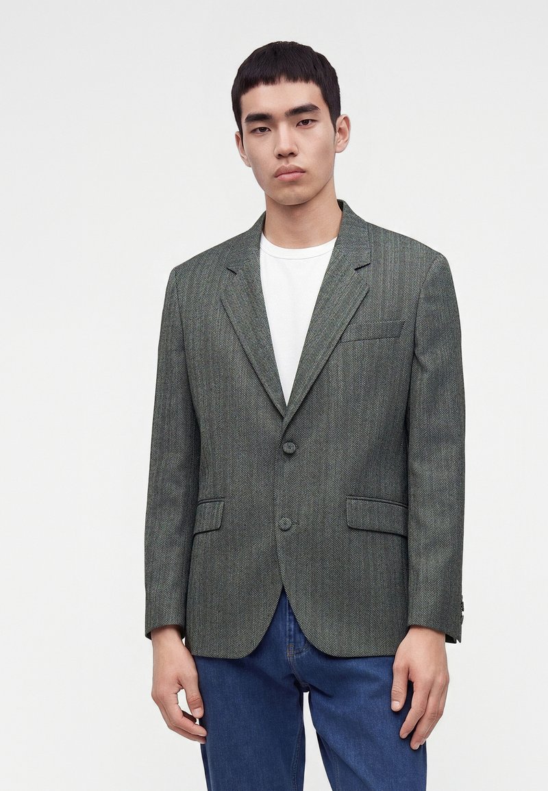 Jeune homme portant un blazer gris à chevrons par-dessus un t-shirt blanc et un jean bleu, debout devant un fond blanc uni.