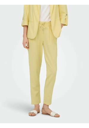 Femme portant un pantalon jaune clair sur mesure avec cordon de serrage, un blazer assorti aux manches retroussées, un top blanc et des sandales tissées blanches.