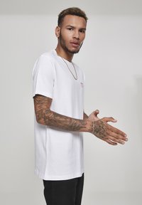 White cotton t-shirt with short sleeves, featuring small red text on the chest. The model has intricate tattoos on both arms.