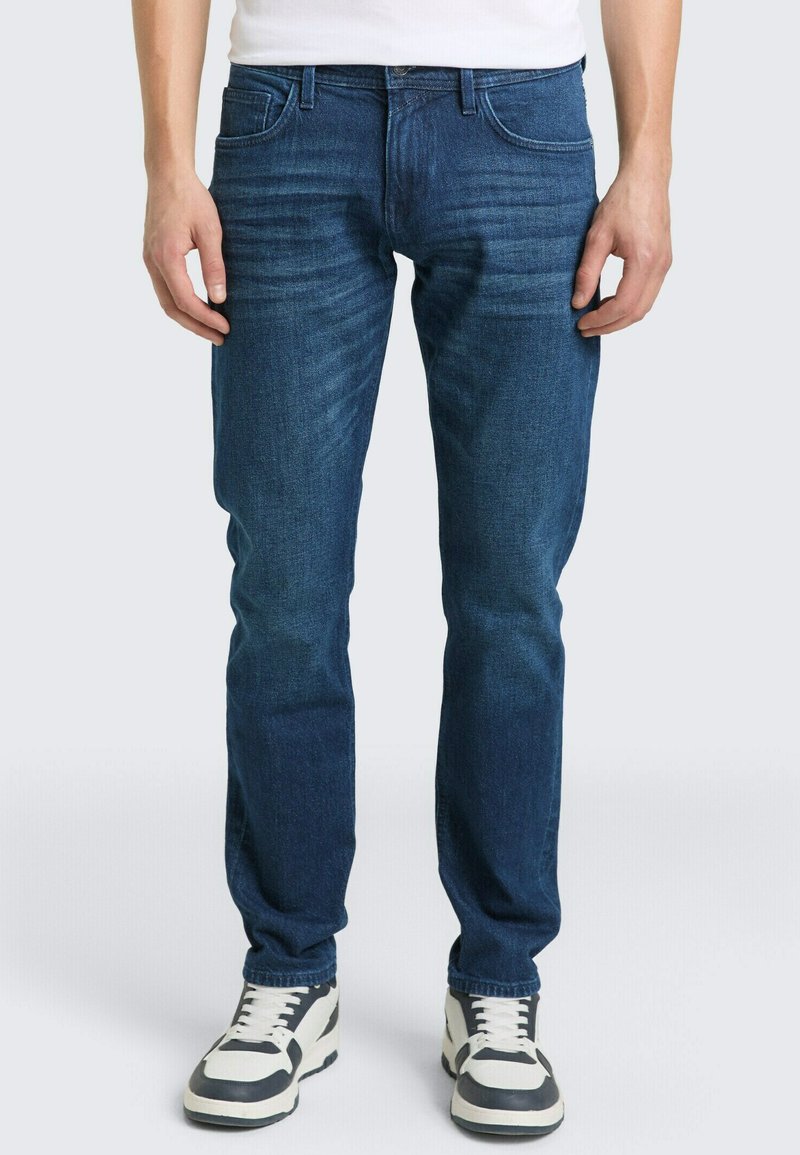 Mężczyzna w dopasowanych niebieskich dżinsach i białych trampkach z czarnymi akcentami, stoi na tle jednolitego jasnego tła.