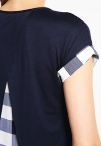 Marinblå kortärmad topp med rutiga uppvikta ärmar och bakpanel. Mjukt tyg med en slät textur samt ett kontrasterande rutmönster.