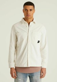 Weißes Button-Up-Hemd mit zwei Brusttaschen, aus strukturiertem Stoff gefertigt. Verfügt über ein schwarzes Logo-Patch und wird über einem hellbraunen Shirt getragen.