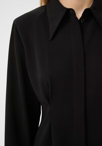 Blouse noire à manches longues avec un col pointu, un tissu lisse, une fermeture à boutons sur le devant et des détails de couture subtils le long de la patte de boutonnage.