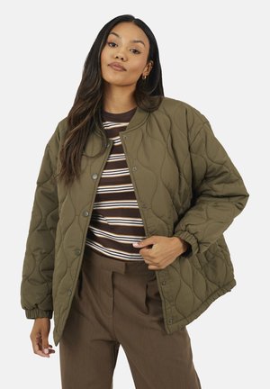 Donna che indossa una giacca trapuntata verde oliva sopra una camicia a righe marroni e pantaloni marroni, in piedi con la mano che sistema la parte frontale della giacca.