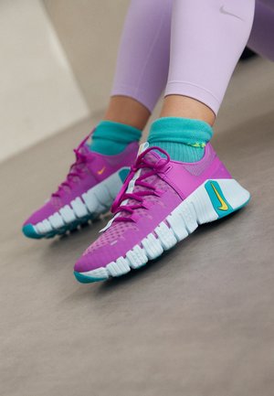 Pies que llevan zapatillas deportivas de color púrpura brillante y turquesa con calcetines turquesas y mallas de color púrpura claro, de pie sobre un suelo liso.