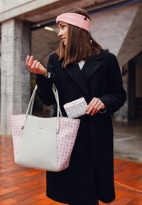 Svetle šedá a růžová taška přes rameno s vzorovaným designem, doplněná odpovídající peněženkou, držena osobou v černém kabátu.