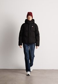 Μαύρο μπουφάν πούφ με ψηλό γιακά, με καπιτονέ υφή και μικρό λογότυπο. Συνδυασμένο με μπλε τζιν και λευκά αθλητικά παπούτσια. Μοντέλο που φοράει μπορντώ σκουφί.