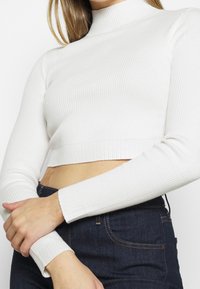 Haut court blanc à côtes avec un col haut, des manches longues et une coupe ajustée. La texture est lisse et extensible. Accompagné d'un pantalon en denim foncé.