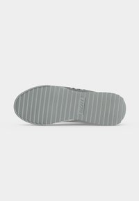 Semelle d'une basket grise avec des rainures horizontales et le logo "DKNY" embossé au centre, sur un fond uni.