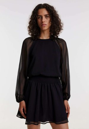 Schwarzes Kleid mit einem durchsichtigen Langarm-Top und einem gerafften Taillenbund. Der Rock ist weit geschnitten und hat einen mehrschichtigen Saum für zusätzliche Textur.