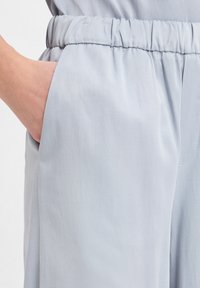 Hand in de zak van lichtblauwe, losse broek met elastische tailleband, waarbij de textuur van de stof en de stiksels zichtbaar zijn.