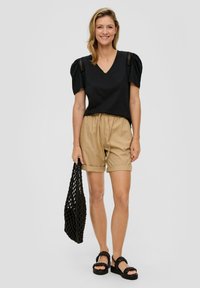 Zwarte v-hals t-shirt met pofmouwen, beige elastische shorts met omslagomslagen, en zwarte geweven tas, gecombineerd met chunky zwarte sandalen.