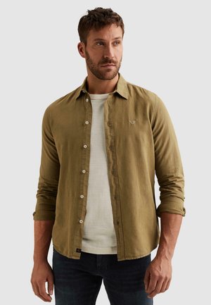 Mand med kort hår og skæg iført en åben olivengrøn skjorte over en hvid tekstureret sweater og mørke jeans, kigger til siden.