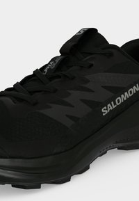 Μαύρο δρομικό παπούτσι Salomon για μονοπάτια με κορδόνια, υφασμένο πέλμα και εμφανές branding "ALPHA GLIDE" και "SALOMON" στο πλάι και στη γλώσσα.