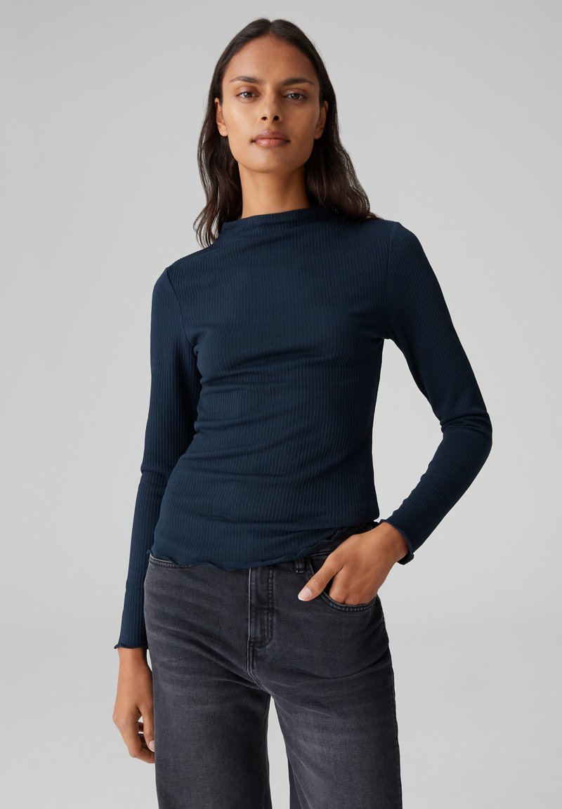 Navy-blaues, geripptes Langarmoberteil mit hohem Kragen und gewelltem Saum, kombiniert mit dunkelgrauen Jeans. Der Stoff hat eine glatte Textur.