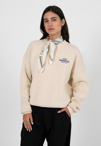 Pull en maille crème à coupe décontractée, avec un logo brodé "PROHIBITED" et une écharpe bleue claire à motifs autour du cou.