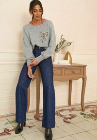 Grauer Pullover mit pinkem Saum und einem Leopardenmuster, kombiniert mit dunkelblaue Schlagjeans und schwarzen Stiefeletten, steht neben einem Holztisch.