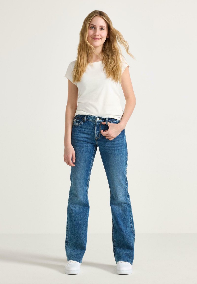 Vit kortärmad t-shirt i kombination med utställda blå denimjeans. Jeansen har en klassisk femfickedesign och är lätt slitna vid fållen.