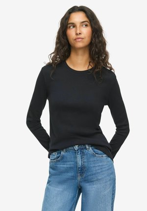 Femme aux longs cheveux bouclés, portant un haut noir à manches longues et un jean bleu, debout les mains derrière le dos, regardant sur le côté.