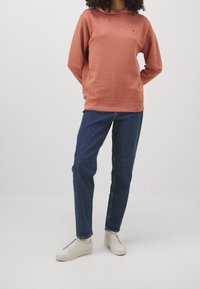 Persona vestida con un sudadera de color óxido, jeans azules y zapatillas blancas, de pie contra un fondo claro y liso.