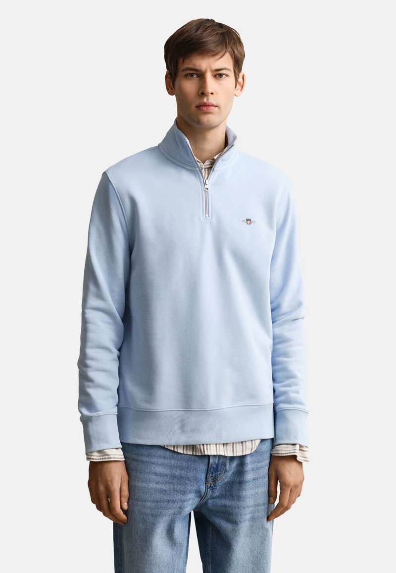 Jeune homme portant un sweat-shirt bleu clair à fermeture éclair quart de zip sur une chemise rayée avec un jean bleu, debout devant un fond blanc uni.