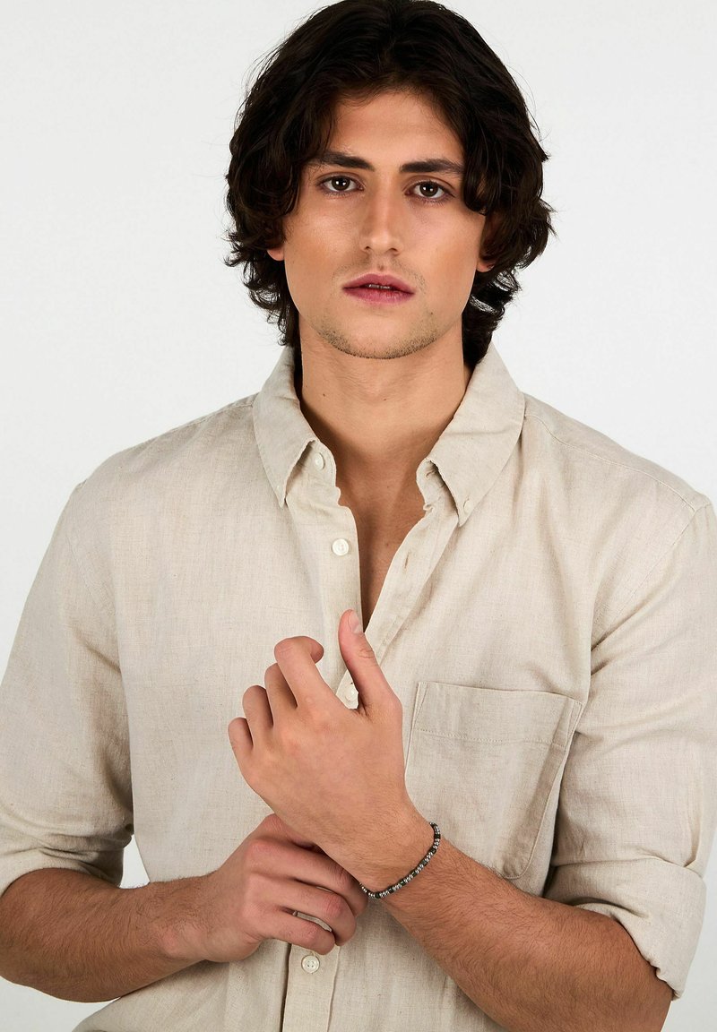 Camicia in lino beige chiaro con colletto allacciato e taschino singolo sul petto. Al polso del modello un braccialetto di perline. Tessuto morbido, vestibilità casual.