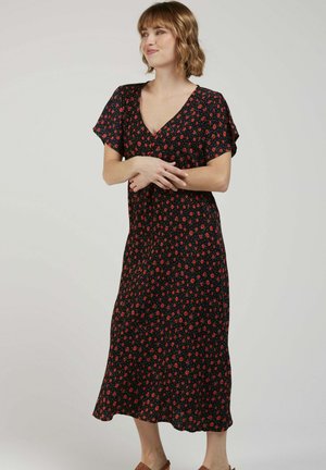 Femme aux cheveux courts et bouclés portant une robe midi noire à imprimé floral rouge, debout les bras croisés et regardant vers la droite.