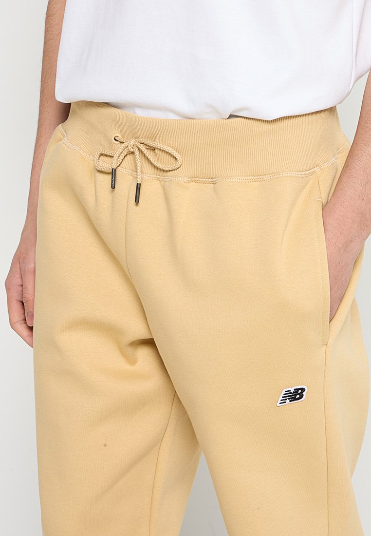Pantalones jogger beige con cintura elástica, cordón y detalle de logo negro. Tejido suave con bolsillos, corte relajado y sin patrones visibles.