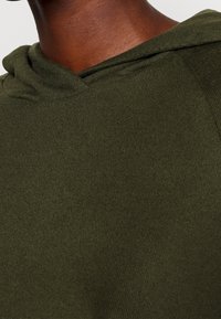 Mörkgrön, ribbad huvtröja i tyg, närbild som visar mjuk textur och detaljer kring huvan, med en svag glans och minimal sömnad.